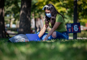 student in mask studying in grass on the quad