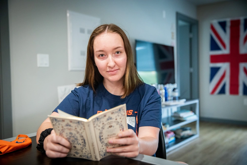 A female student is reading a book with a TV and a UK flag in the background.